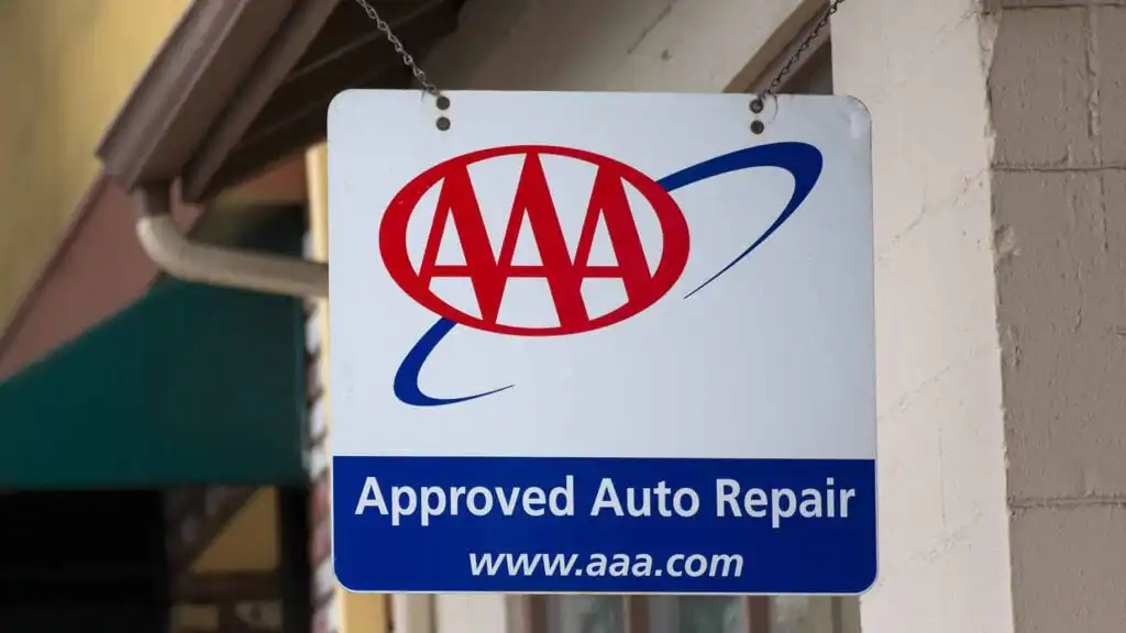 A sign hanging outside a building reads AAA Approved Auto Repair with the AAA logo and website address www.aaa.com on a white and blue background.
