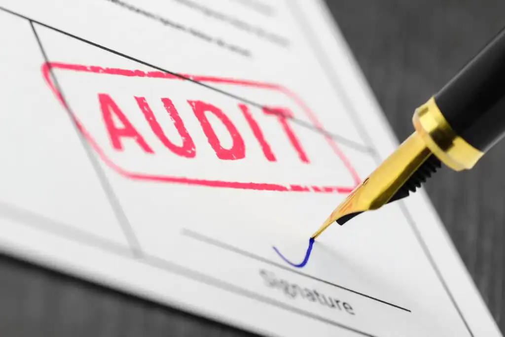 A close-up of a document stamped with the word AUDIT in red ink, with a fountain pen signing the signature line at the bottom of the page.