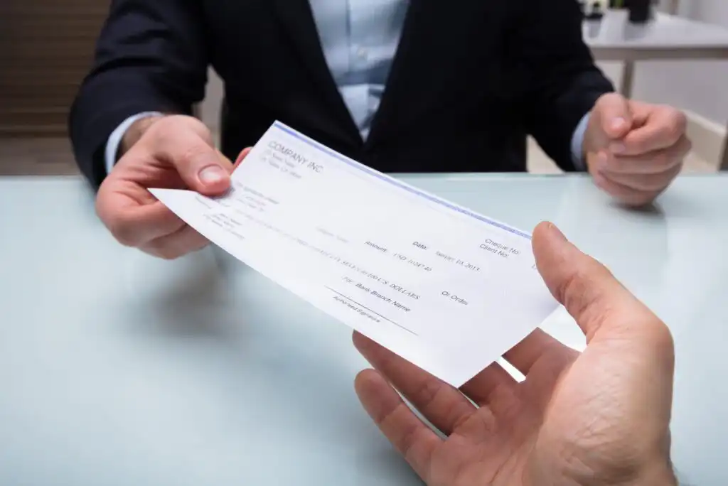 A person in a suit hands a check to another person across a glass table, with only their hands and part of their torsos visible.