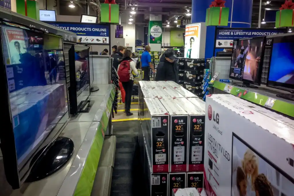 Shoppers browse aisles filled with boxed LG televisions and display screens in an electronics store, with signs, bright lights, and holiday gift decorations visible throughout the busy scene.