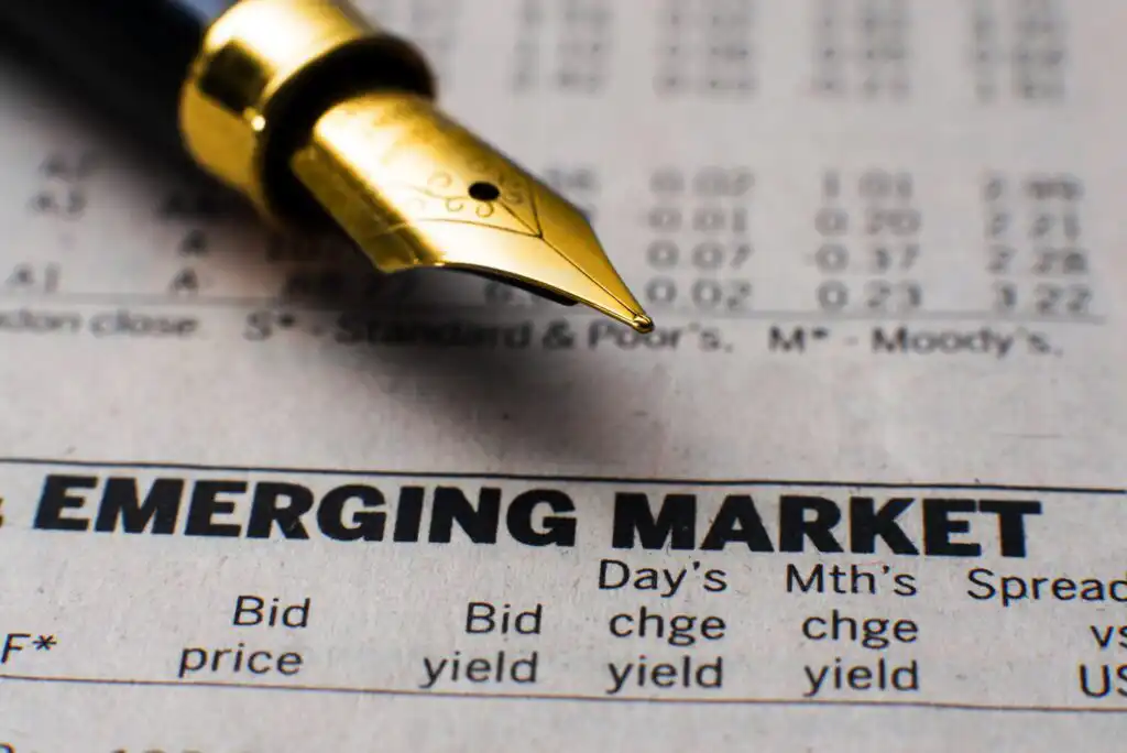 A gold fountain pen rests on a financial newspaper, highlighting the headline EMERGING MARKET above columns for bid price, yield, and daily change.
