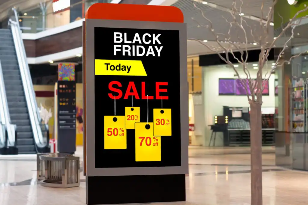 A digital display in a shopping mall advertises a Black Friday sale with yellow and red tags showing 50%, 70%, 30%, and 20% off. The background features shops, an escalator, and a leafless decorative tree.