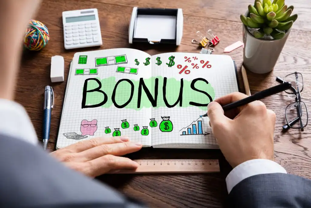 A person writes BONUS in a notebook with colorful drawings of money, charts, and financial symbols, surrounded by office supplies, a calculator, and a potted plant on a wooden desk.