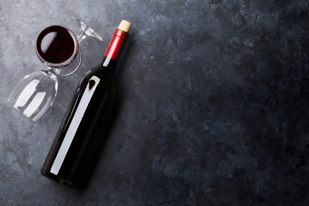 A bottle of red wine with a red foil top lies on a dark stone surface next to two wine glasses, one upright filled with wine and the other empty and tipped over.