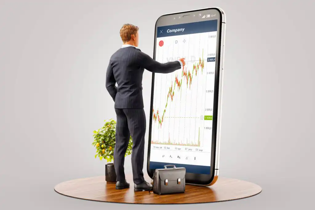 A businessman in a suit points at a large smartphone screen displaying a rising stock chart. He stands on a wooden surface beside a plant and a briefcase, symbolizing financial or stock market analysis.