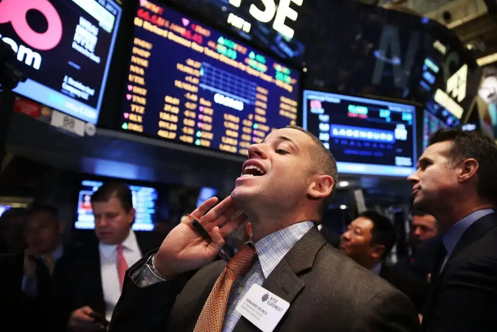 A stock trader on the busy floor of the New York Stock Exchange gestures and shouts, surrounded by colleagues and large electronic boards displaying stock prices and market data.