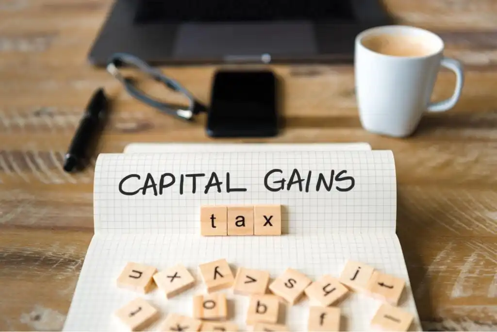 A notepad with CAPITAL GAINS written on it and Scrabble tiles spelling tax, surrounded by more tiles. In the background are a laptop, glasses, smartphone, pen, and a cup of coffee on a wooden table.