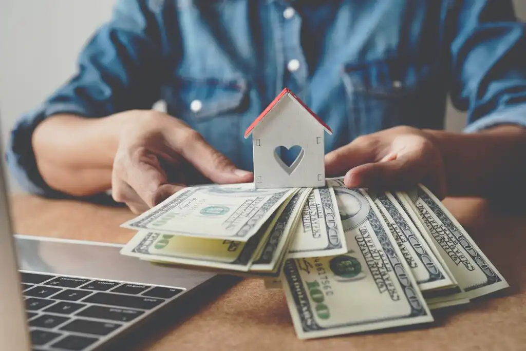 A person holds a small house model over several spread-out US hundred-dollar bills next to a laptop, symbolizing real estate investment or home buying.