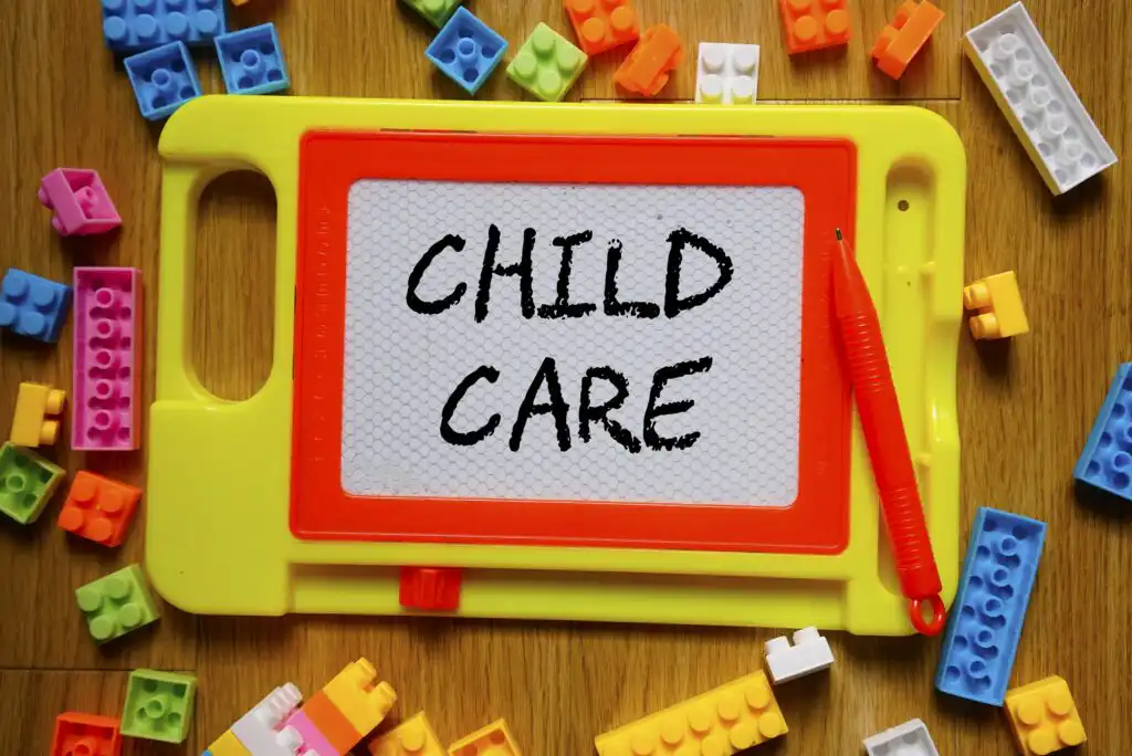 A yellow and red magnetic drawing board with “CHILD CARE” written on it, surrounded by colorful toy building blocks and a red magnetic stylus, all placed on a wooden surface.