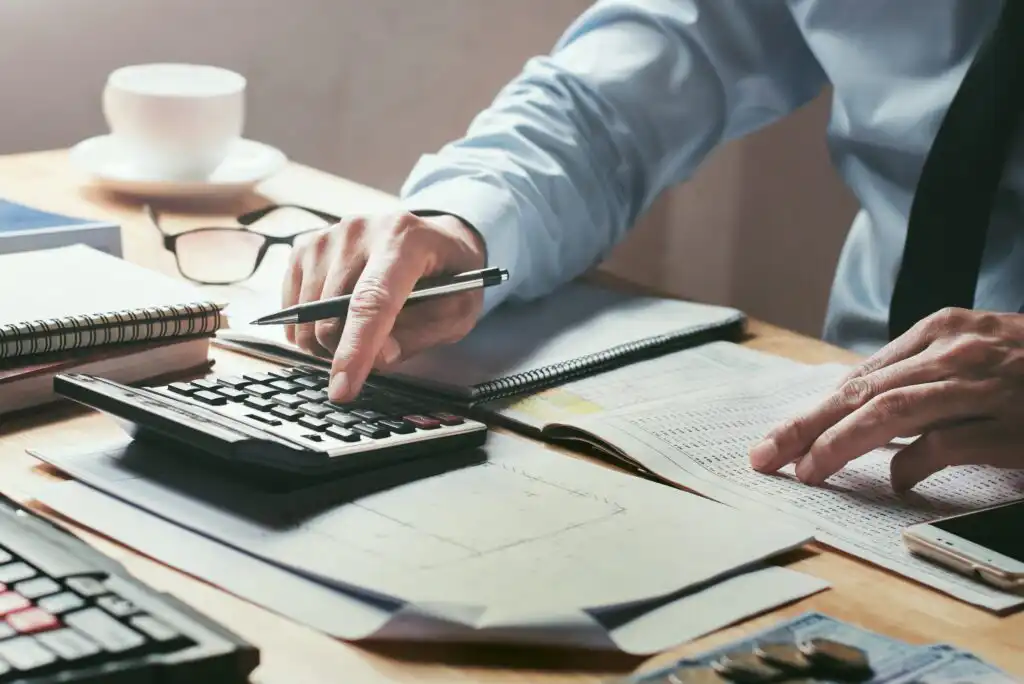 A person in business attire uses a calculator and reviews financial documents at a cluttered desk with papers, a notebook, glasses, and a cup in the background.