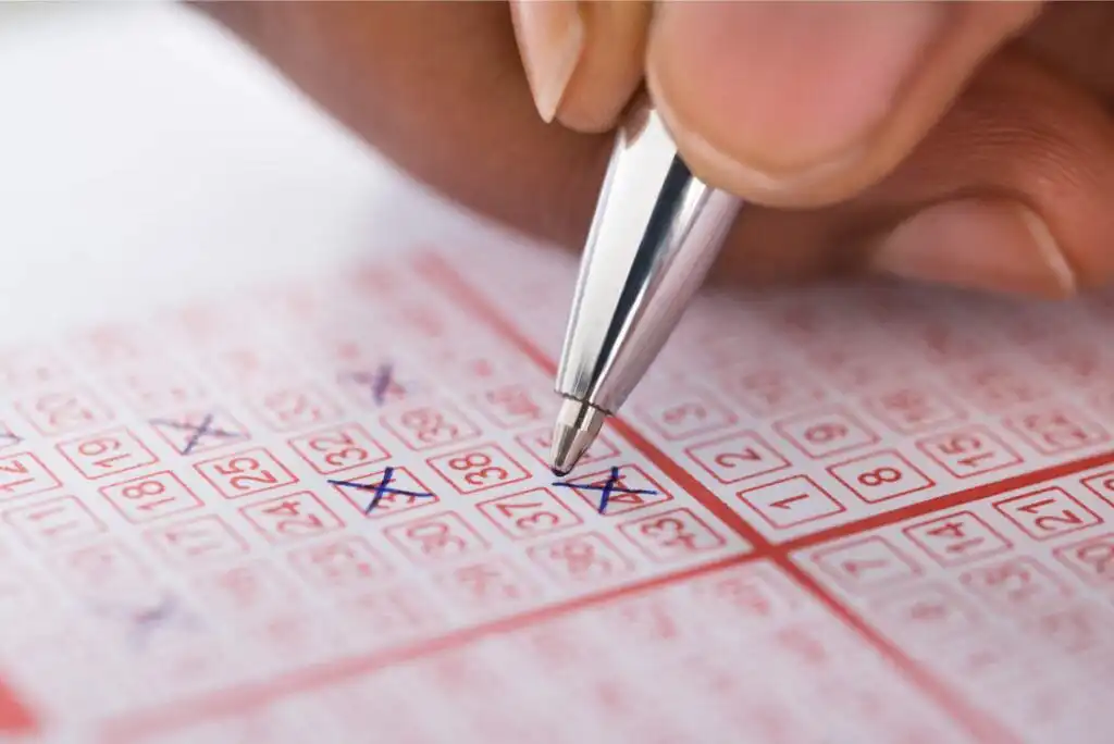 A close-up of a hand holding a pen and marking numbers on a lottery ticket, with several numbers already crossed out in blue ink.