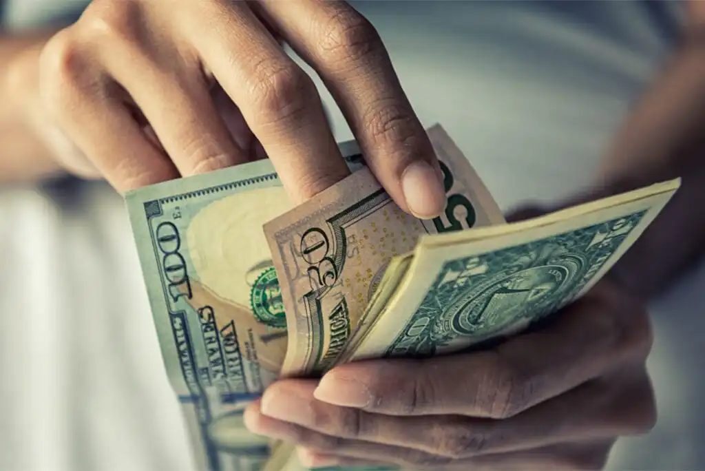 A person’s hand is holding and sorting several U.S. dollar bills, including a $100 bill, a $20 bill, and a $1 bill. The background is blurred.