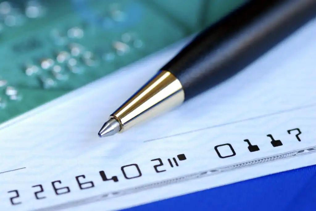 A close-up of a pen resting on a personal check, showing the checks routing and account numbers in magnetic ink character recognition (MICR) font. A blurred circuit board is visible in the background.