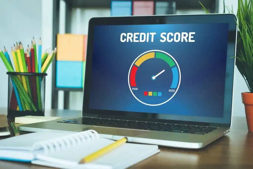 A laptop on a desk displays a credit score gauge ranging from poor to good. The workspace includes a notebook, pen, potted plant, and shelves with colorful folders in the background.