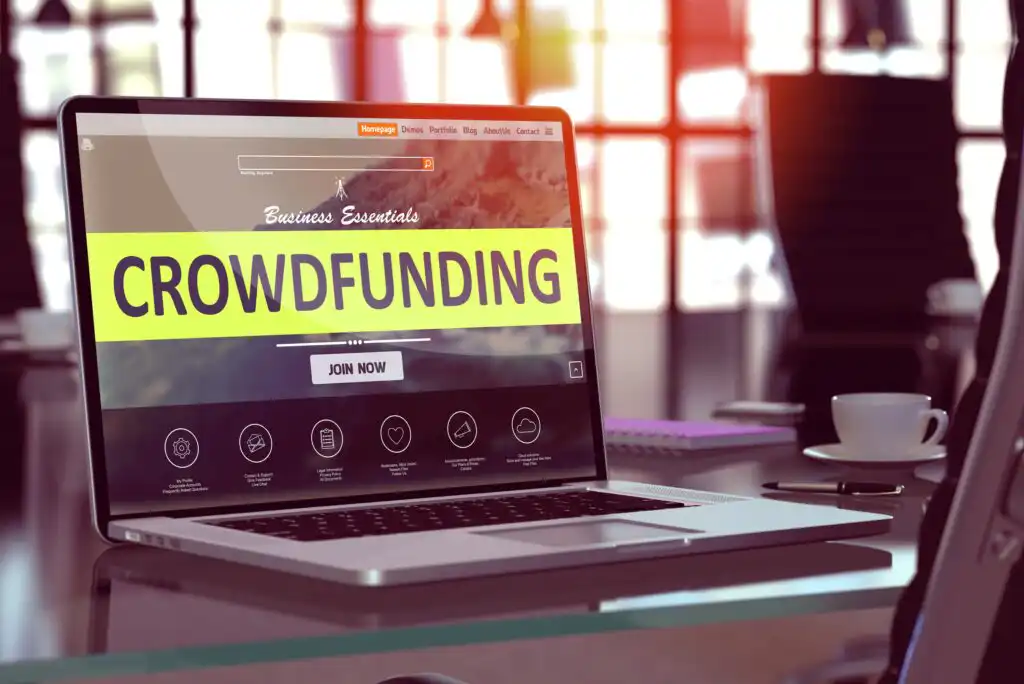 A laptop on a glass desk displays a website with a large yellow banner reading CROWDFUNDING and a Join Now button. The background shows an office with large windows and blurred light.
