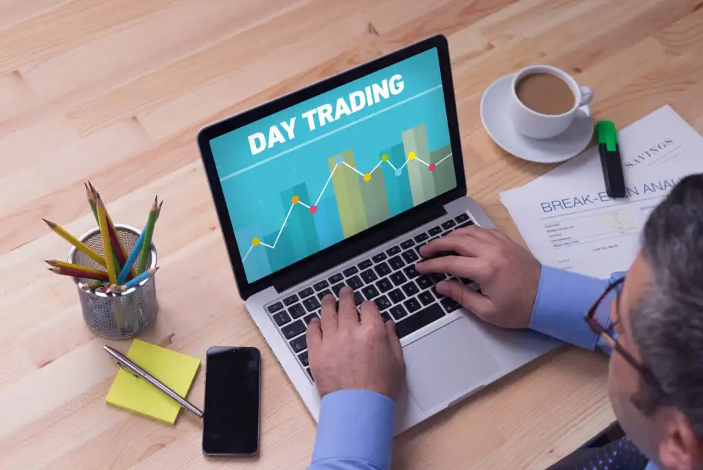 A person using a laptop displaying a screen with the words DAY TRADING and a financial chart. Office supplies, a smartphone, paper, and a cup of coffee are on the wooden desk.