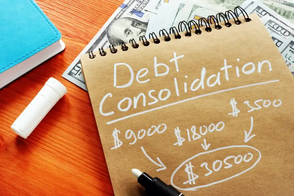 A notebook with Debt Consolidation written on it, showing various amounts totaling $30,500, lies on a wooden table with a marker, cash, and a blue notebook nearby.