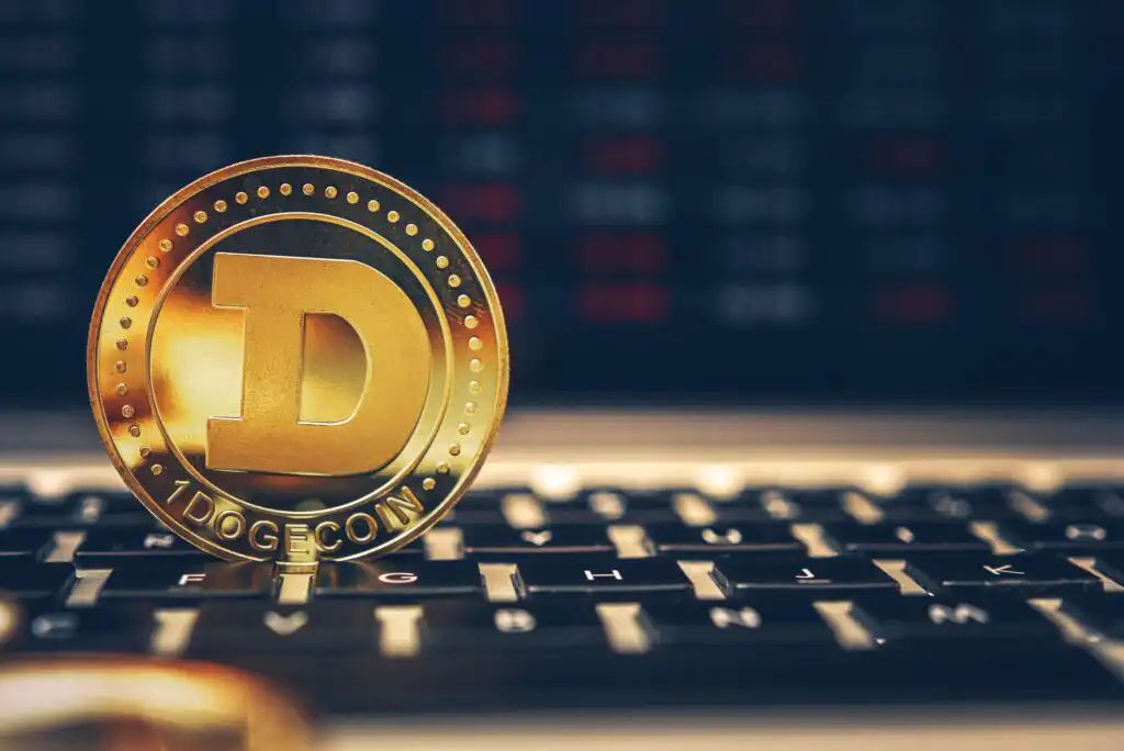 A gold Dogecoin token standing upright on a computer keyboard, with a blurred screen displaying red and black data in the background.