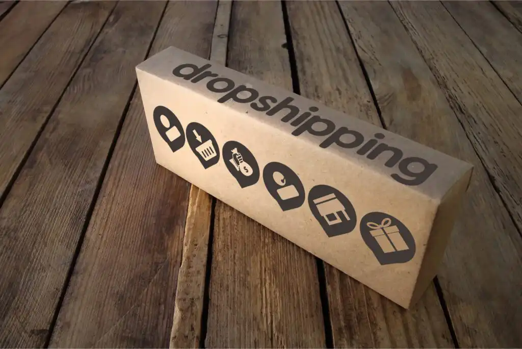 A cardboard box labeled dropshipping sits on a wooden floor, featuring black icons representing various stages of shipping and delivery, including a truck, location pin, store, box, and gift.