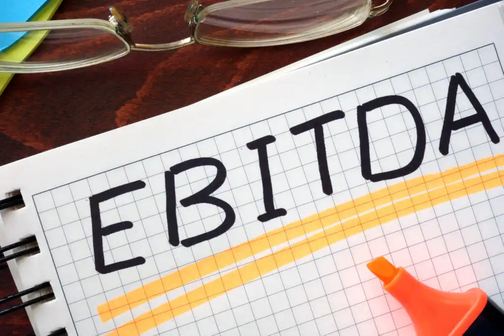 A notebook with the word EBITDA written in bold letters on a grid page, highlighted with orange marker, with a pair of eyeglasses and part of a highlighter visible nearby.