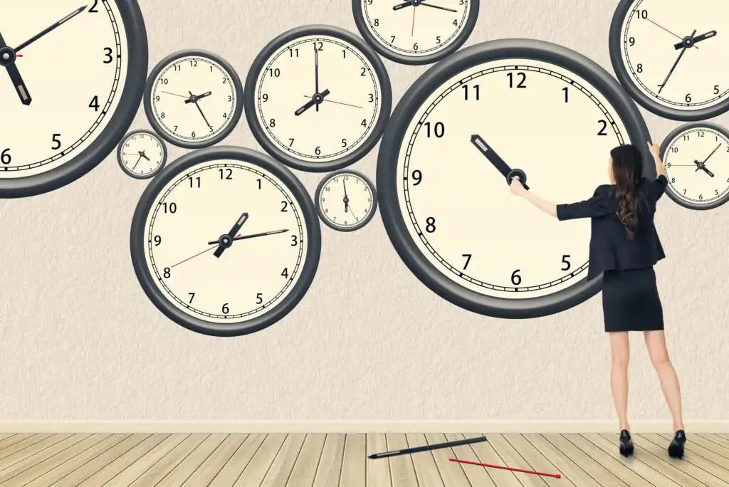 A woman in business attire stands on tiptoes adjusting the hands of a large wall clock among several other clocks on a white wall, with clock hands scattered on the wooden floor.