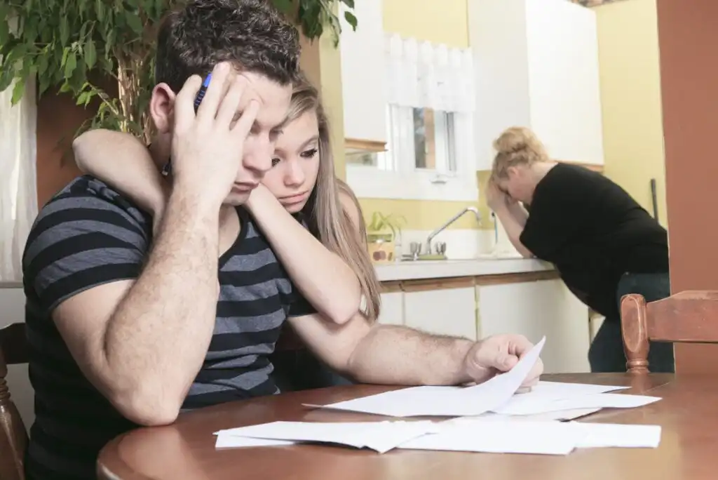 A young couple sits at a table looking worried as they review documents; the woman hugs the man, and another woman stands in the background, visibly upset with her head in her hand.