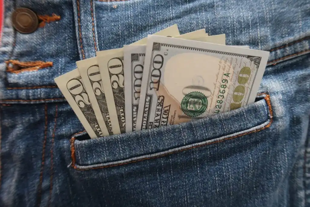 Several U.S. dollar bills, including $100 and $20 denominations, are sticking out of the front pocket of blue denim jeans.