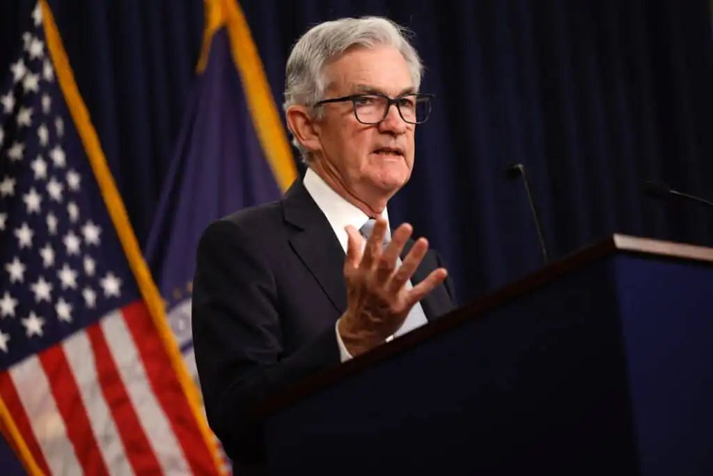 A man in a suit and glasses speaks at a podium with an American flag and another flag in the background. He gestures with one hand, appearing to address an audience.