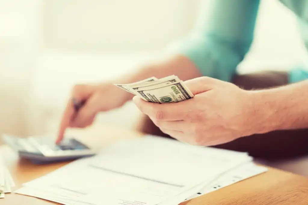 A person holds several U.S. hundred-dollar bills in one hand while using the other hand to press buttons on a calculator, with documents and papers on a table in front.