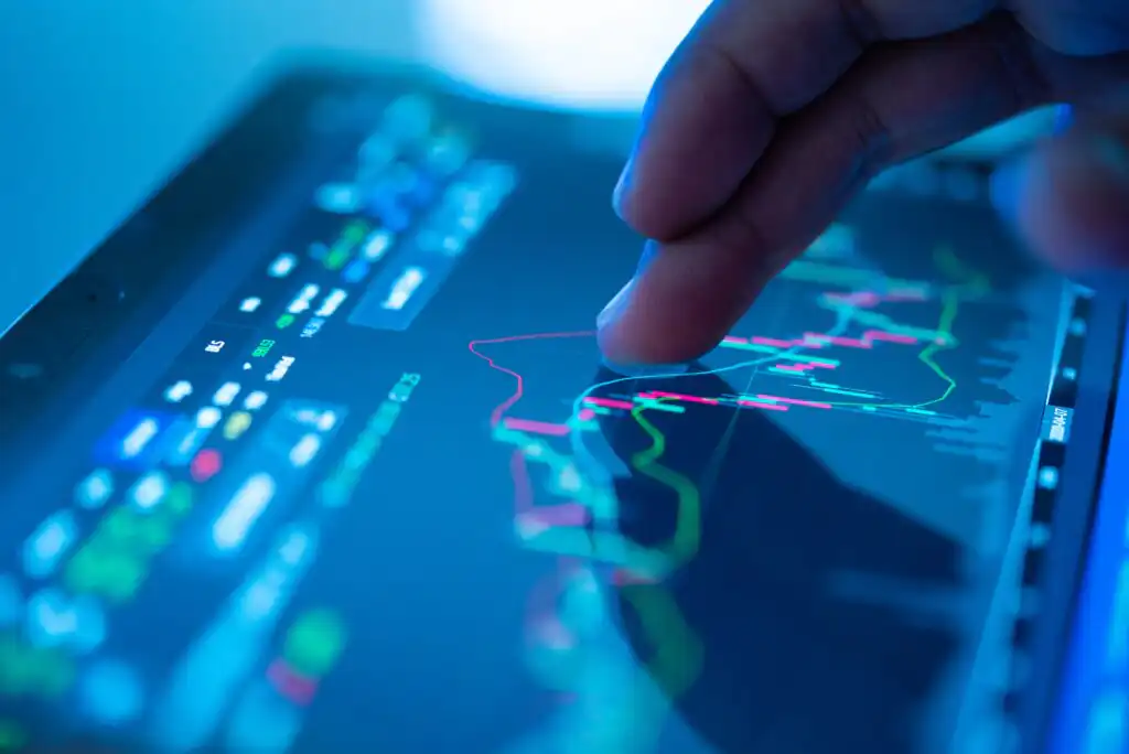 A hand interacts with a tablet displaying colorful financial graphs and data, including green and red lines representing stock market trends, against a blue-lit background.