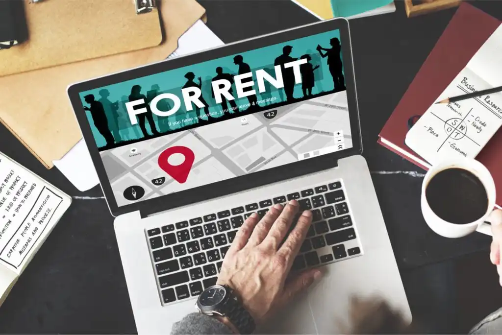 A person using a laptop displaying a “For Rent” web page with a map and location pin. A cup of coffee, notebook, pen, and folders are visible on the desk.