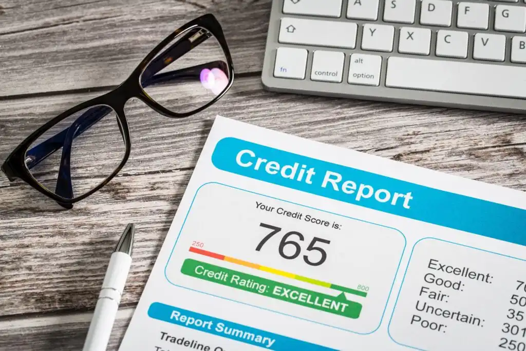 A close-up of a credit report showing a credit score of 765 rated as excellent, with a keyboard, eyeglasses, and pen on a wooden desk.