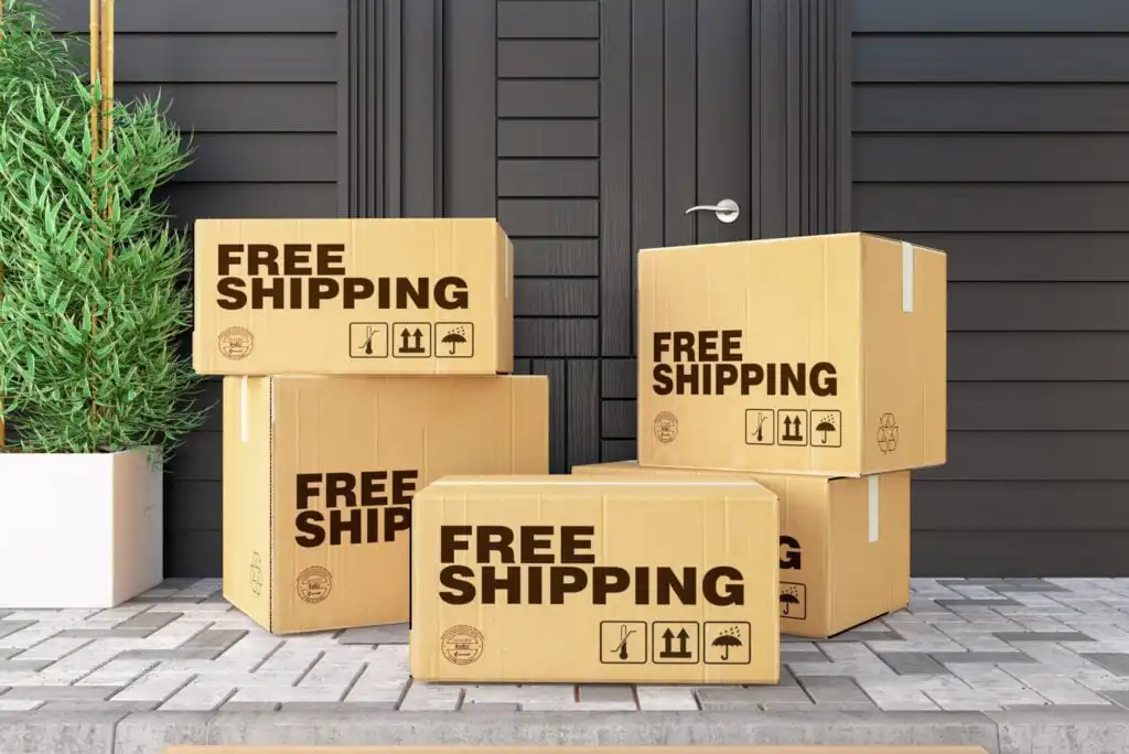 Several cardboard boxes labeled FREE SHIPPING are stacked in front of a modern black door with a silver handle, next to a green plant on a paved doorstep.
