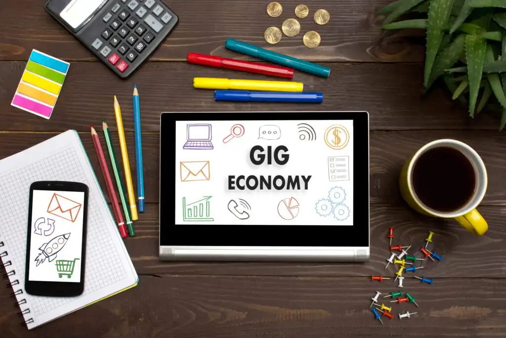 A tablet on a wooden desk displays “Gig Economy” with icons. Surrounding items include colored pencils, a calculator, coins, a notepad with a phone, a cup of coffee, push pins, and a plant.