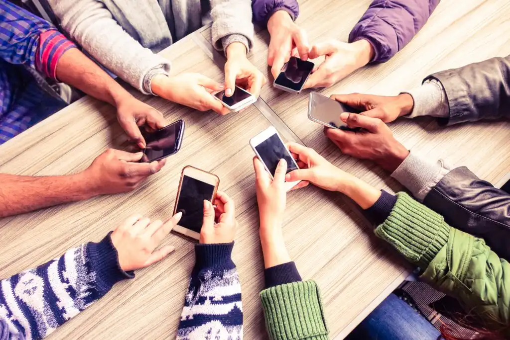 A group of people sitting around a table, each holding and using a smartphone, with only their hands and arms visible. The table is wooden and the scene shows various sleeves and jackets.