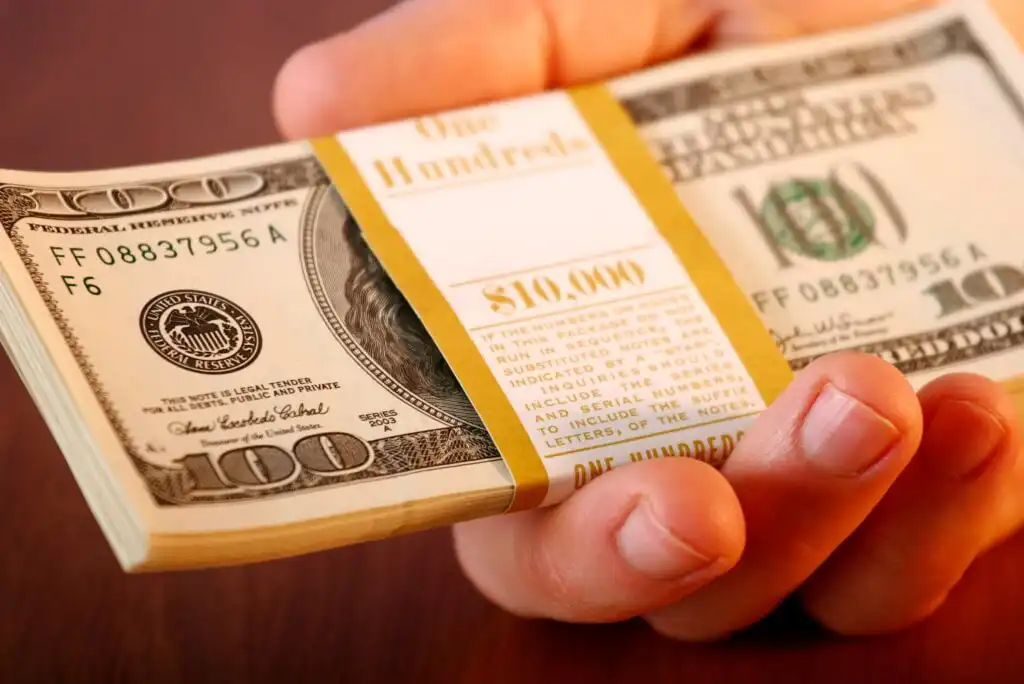 A hand holding a thick stack of $100 US bills bound with a paper band labeled $10,000 on a wooden surface.