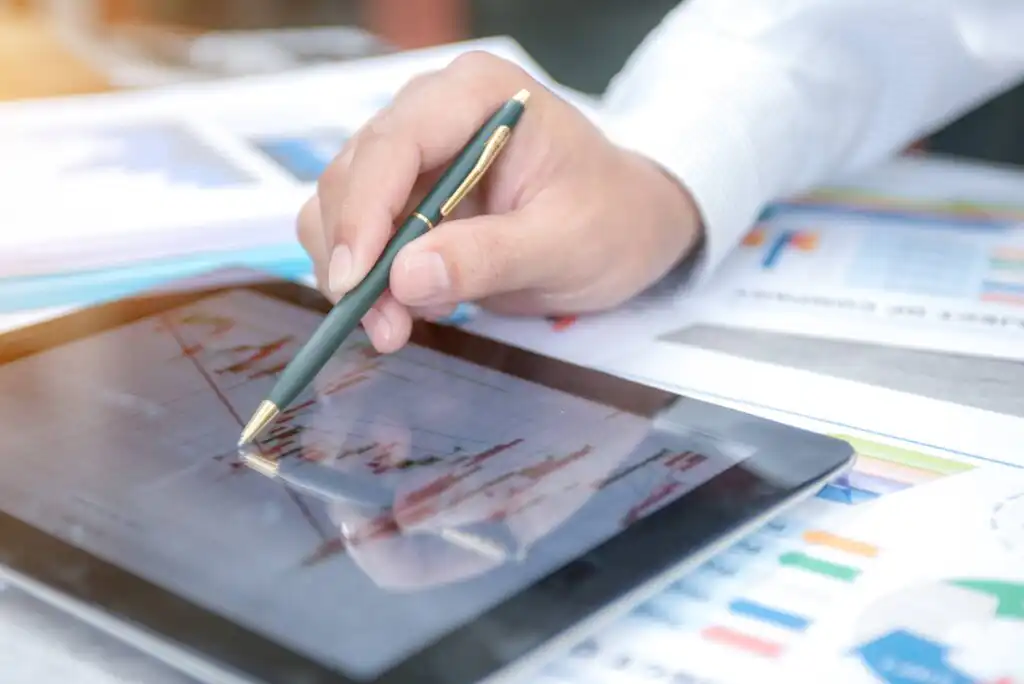A person’s hand holding a pen points at a financial graph displayed on a tablet, with colorful charts and documents spread out on the table in the background.