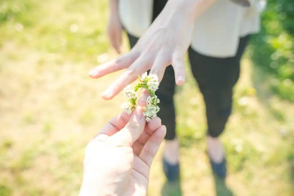 A person puts a delicate flower ring made of small white blossoms on another persons finger outdoors in bright sunlight. The background is grassy and slightly blurred.