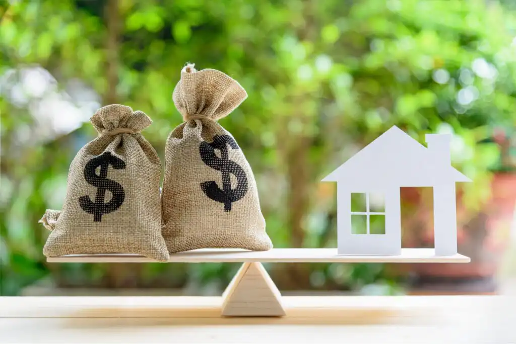 Two burlap money bags with dollar signs balance on one side of a wooden seesaw, while a white paper house balances on the other side. The background is blurred greenery.