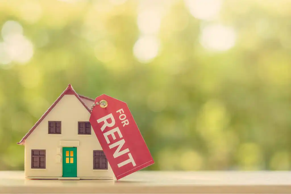 A small model house with a green door has a red tag labeled FOR RENT attached to its roof, set against a blurred, sunlit, green background.