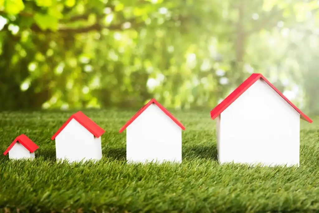 Four small model houses with red roofs, arranged in increasing size from left to right, sit on green grass with a blurred, sunlit background of trees.