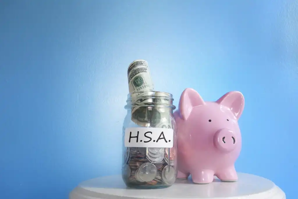A glass jar labeled H.S.A. filled with coins and cash stands next to a pink piggy bank against a light blue background, both placed on a white surface.