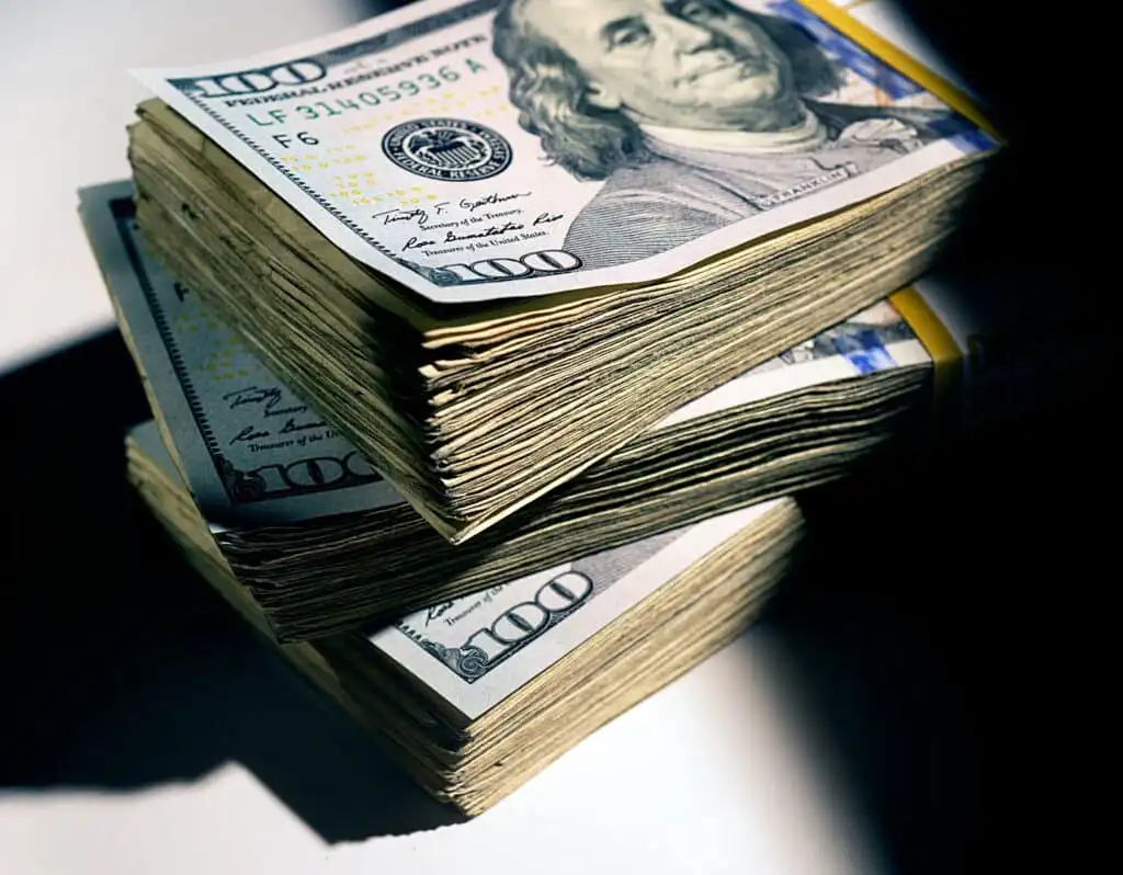 Two thick stacks of 100-dollar bills are placed on a flat surface, with the top bill in each stack showing Benjamin Franklins portrait. Bright light casts sharp shadows on the money.