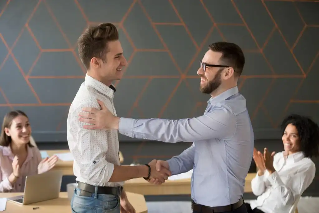 Two men standing and shaking hands while smiling at each other in an office. Two colleagues sitting at desks in the background are applauding. All appear happy and professionally dressed.