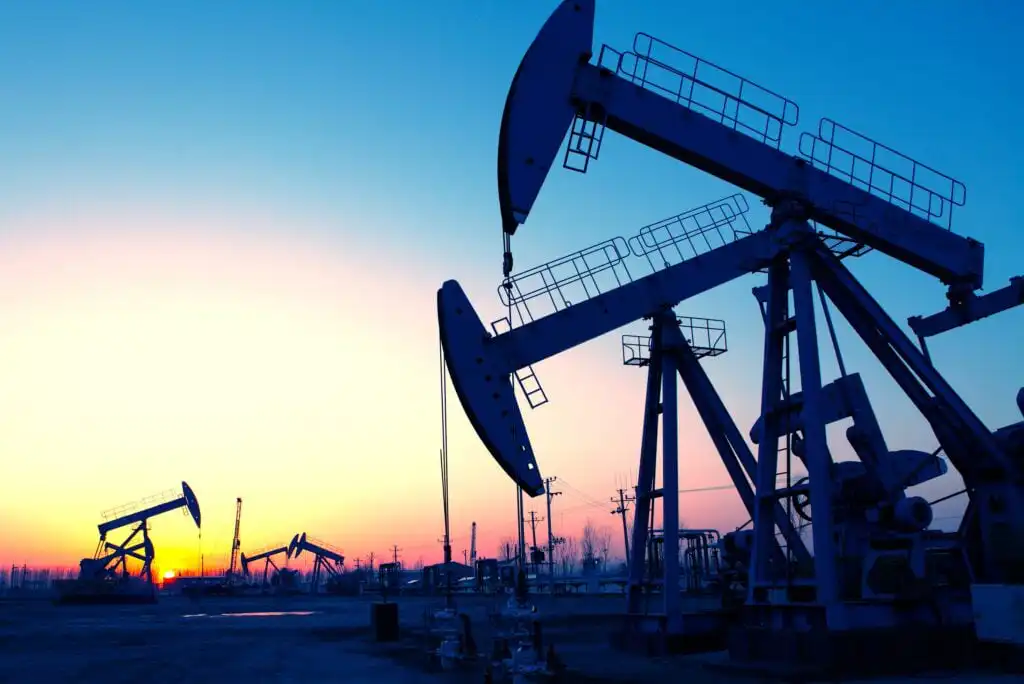 Oil pumpjacks silhouetted against a colorful sky at sunrise or sunset, with several pumps scattered across a flat landscape, power lines visible in the background.