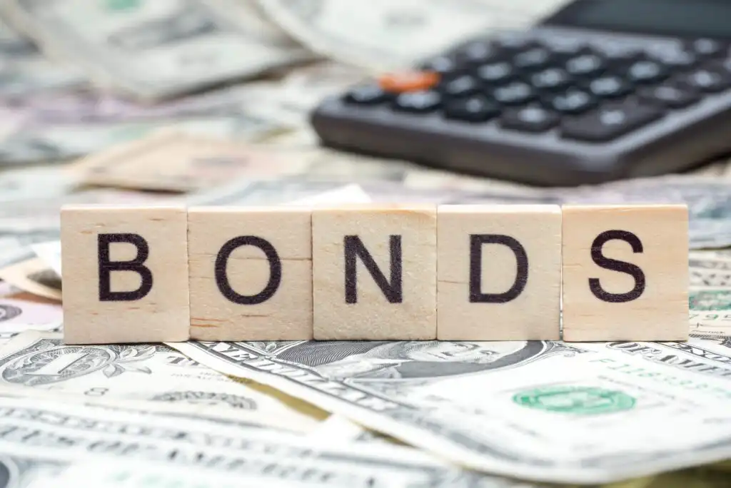 Wooden blocks spelling BONDS sit on top of scattered U.S. dollar bills, with a blurry calculator in the background, symbolizing finance and investments.