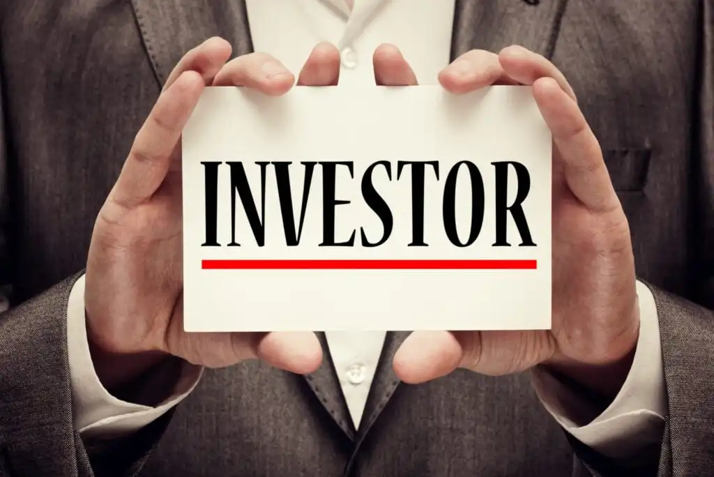 A person in a suit holds a white card with the word INVESTOR in bold black letters, underlined in red.