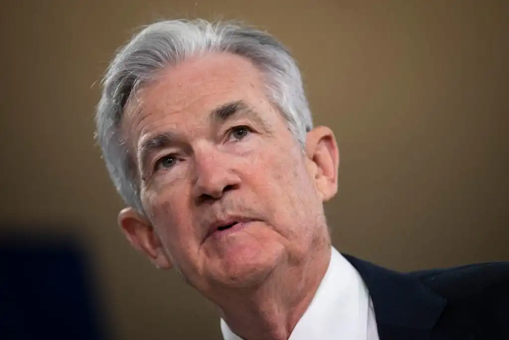 An older man with gray hair wearing a dark suit and white shirt looks slightly upward with a serious expression against a blurred background.