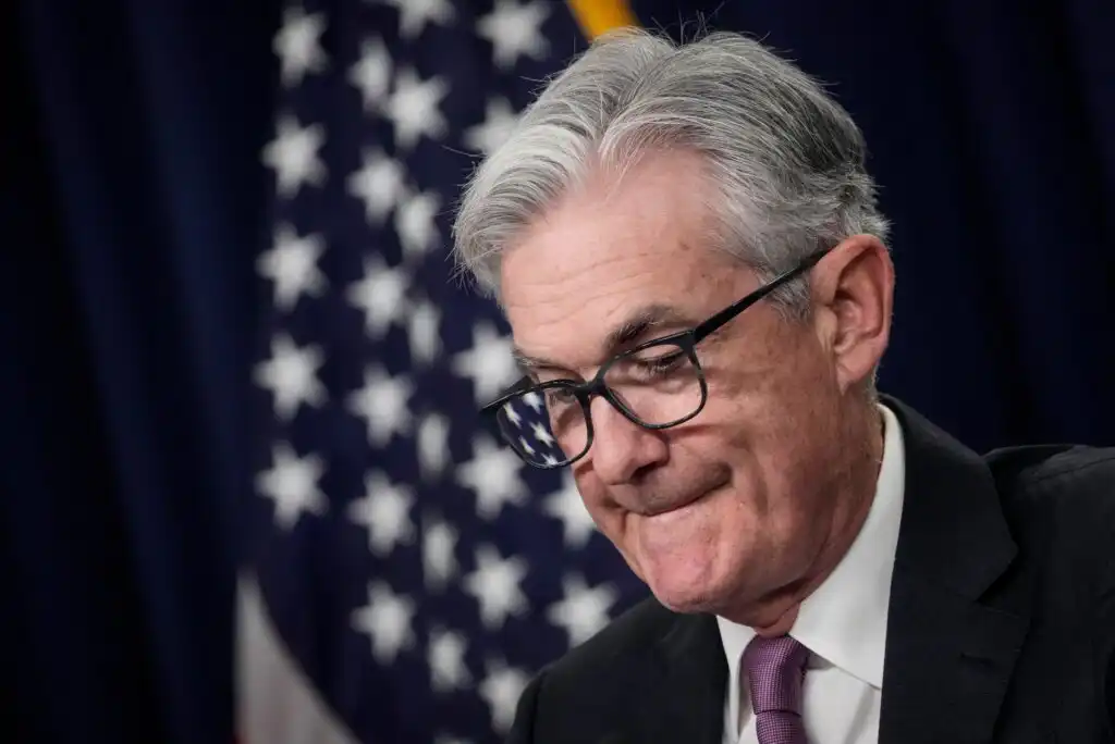 A man with gray hair and glasses, wearing a suit and tie, stands in front of a blurry American flag, looking downward with a serious expression.