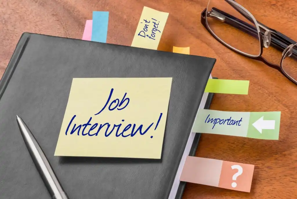 A notebook with colorful sticky tabs and a pen rests on a wooden desk. A yellow sticky note on the cover reads Job Interview! in blue handwriting, and tabs are labeled Dont forget!, Important, and ?.